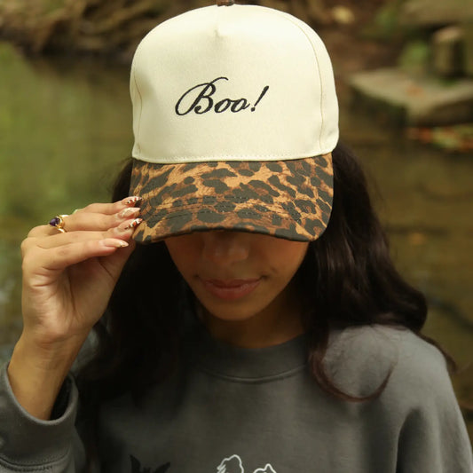 Person wearing a cap with 'Boo!' text and leopard print brim, outdoors.