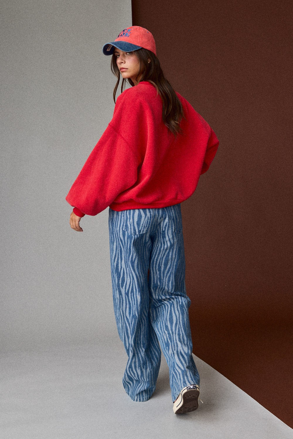 Person wearing a red sweater and blue jeans with a red cap against a gradient wall.