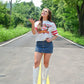 Woman tossing a football in the air while smiling, wearing a cropped white Kansas City Chiefs tee, a pinstriped denim mini skirt, and white embroidered cowboy boots, standing on a sunny tree-lined road.