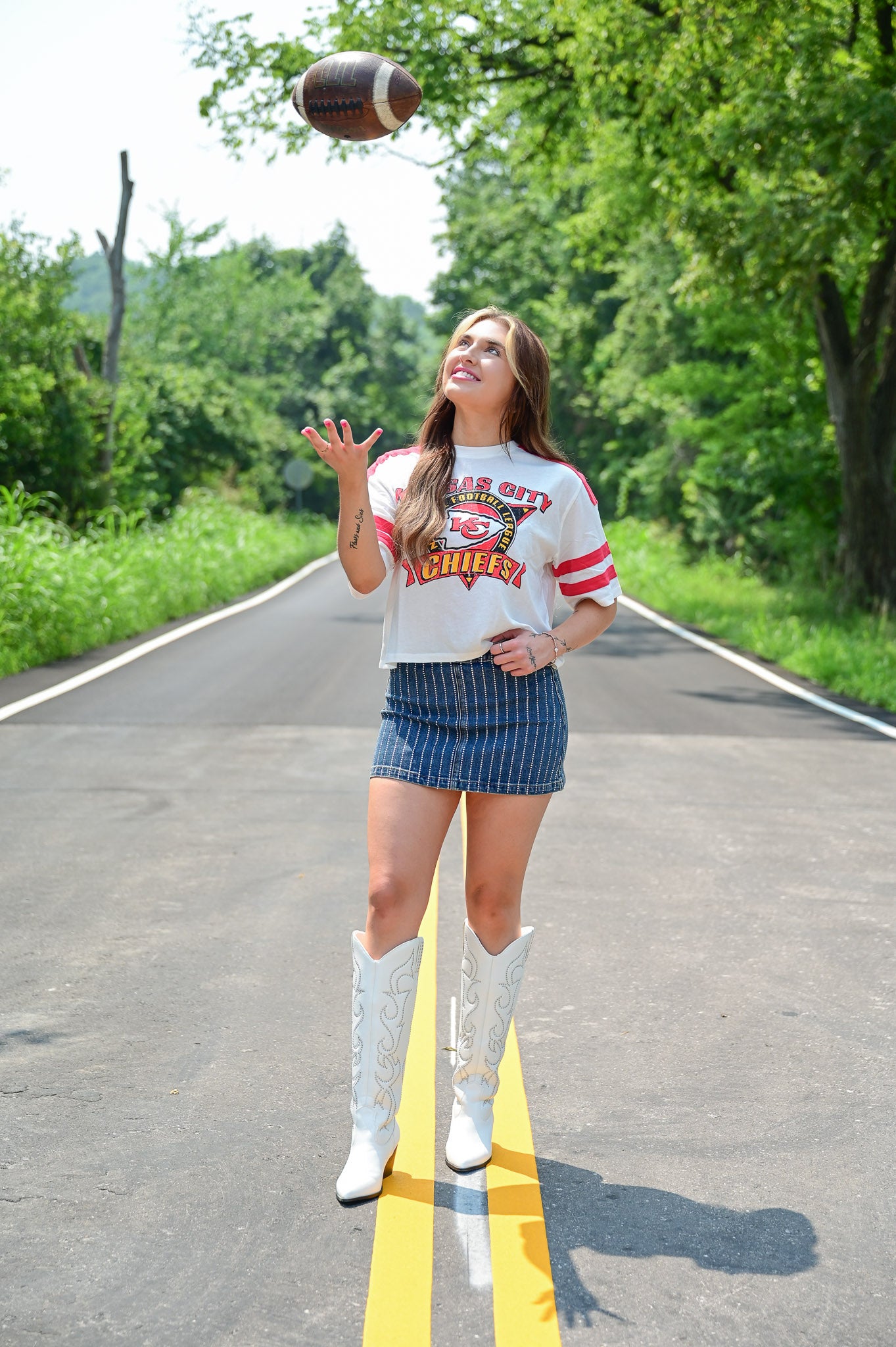 Woman tossing a football in the air while smiling, wearing a cropped white Kansas City Chiefs tee, a pinstriped denim mini skirt, and white embroidered cowboy boots, standing on a sunny tree-lined road.
