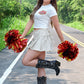 Woman smiling while holding red and yellow pom-poms in both hands, wearing a white Kansas City Chiefs crop top, cream wrap mini skirt, and black embroidered cowboy boots, posing playfully on a road surrounded by greenery.
