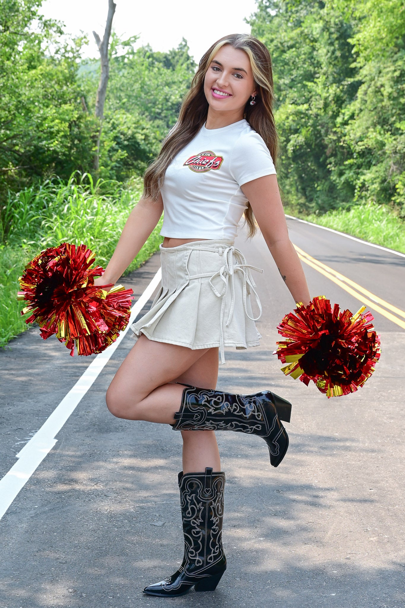 Woman smiling while holding red and yellow pom-poms in both hands, wearing a white Kansas City Chiefs crop top, cream wrap mini skirt, and black embroidered cowboy boots, posing playfully on a road surrounded by greenery.