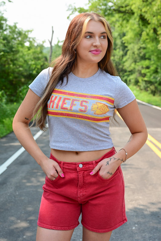 Woman standing on a road wearing a fitted gray crop top with a vintage-style Kansas City Chiefs logo and red high-waisted shorts, with greenery in the background.