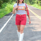 Woman walking down a road in a gray Kansas City Chiefs crop top, red shorts, white embroidered cowboy boots, and a red shoulder bag, smiling with trees lining the road.