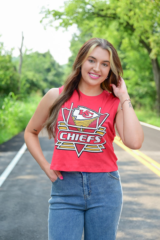 Woman smiling and posing outdoors in a red Kansas City Chiefs sleeveless crop top and high-waisted blue jeans, standing on a road with greenery in the background.