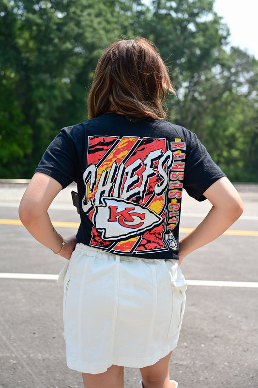 Woman standing outdoors wearing a black Kansas City Chiefs graphic tee and a white high-waisted utility skirt, with her back to the camera and hands on her hips.