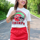 Woman smiling while wearing a cropped white Kansas City Chiefs t-shirt with a football helmet graphic and a red athletic mini skirt with side ties, standing on a road lined with trees.