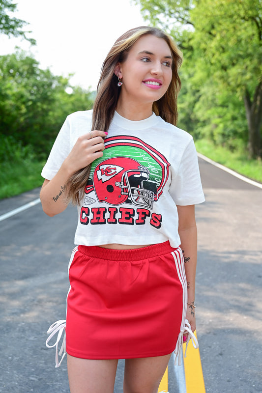 Woman smiling while wearing a cropped white Kansas City Chiefs t-shirt with a football helmet graphic and a red athletic mini skirt with side ties, standing on a road lined with trees.