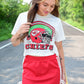 Woman looking down and smiling in a white Chiefs crop top featuring a red helmet and green field graphic, paired with a red skirt with white stripes and side ties, standing on a tree-lined road.