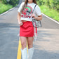 Woman posing confidently on a road in a cropped Chiefs helmet tee, red athletic mini skirt with white stripes, white cowboy boots, and a clear crossbody purse, with greenery in the background.