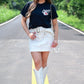 Woman standing confidently on a road wearing a black Kansas City Chiefs t-shirt, white mini skirt with pockets, studded belt, white embroidered cowboy boots, and a clear crossbody purse, with greenery in the background.