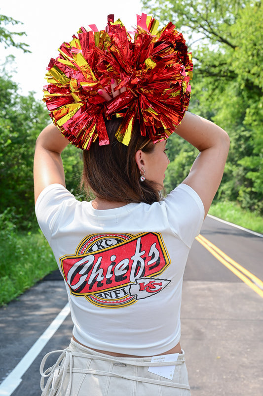 Woman holding red and yellow metallic pom-poms above her head, wearing a white cropped Kansas City Chiefs graphic tee and a cream mini skirt, with her back to the camera on a tree-lined road.