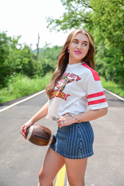 Woman posing on a road holding a football, wearing a cropped white Kansas City Chiefs t-shirt with red stripes on the sleeves and a high-waisted, pinstriped denim mini skirt, surrounded by greenery.