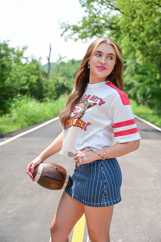 Woman posing on a road holding a football, wearing a cropped white Kansas City Chiefs t-shirt with red stripes on the sleeves and a high-waisted, pinstriped denim mini skirt, surrounded by greenery.